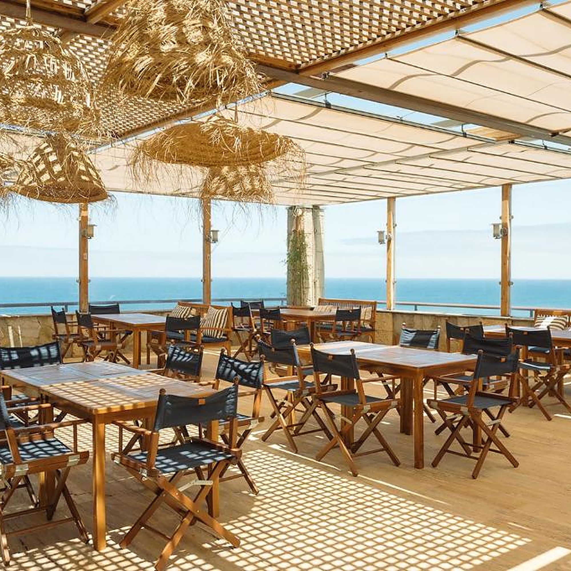An outdoor seating area with tables and chairs, a thatched ceiling, and a view of the sea.