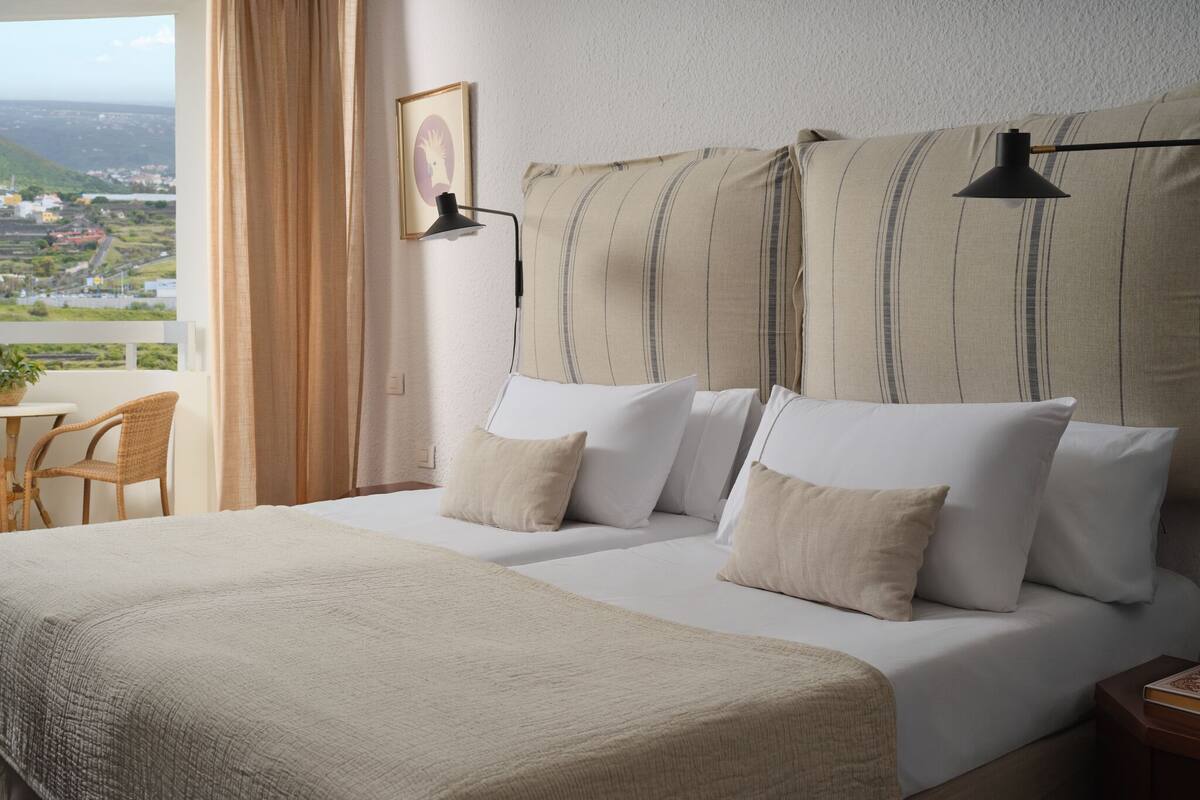 Hotellihuone, jossa on sänky, näköala kaupunkiin, pöytä tuoleineen ja lamppu.