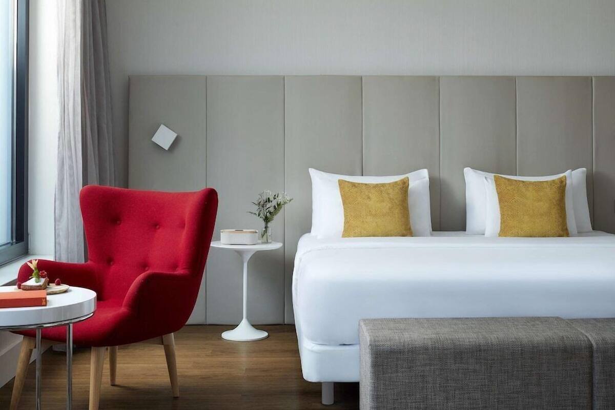 Ein Hotelzimmer mit einem Bett, einem roten Sessel, einem kleinen Nachttisch und einem grauen Hocker.