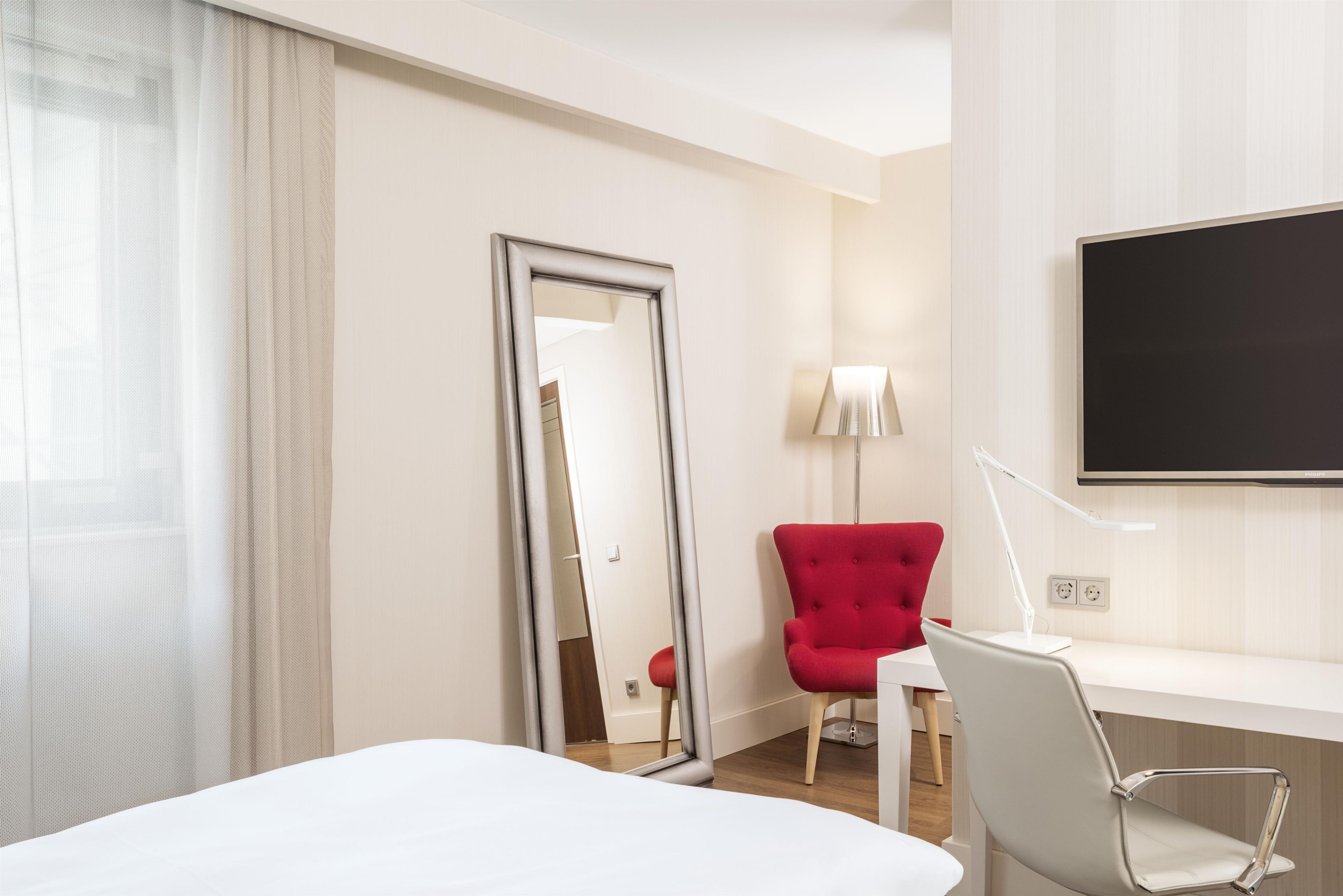 Habitación de hotel con cama, una silla roja, escritorio con lámpara, televisor de pantalla plana y espejo.