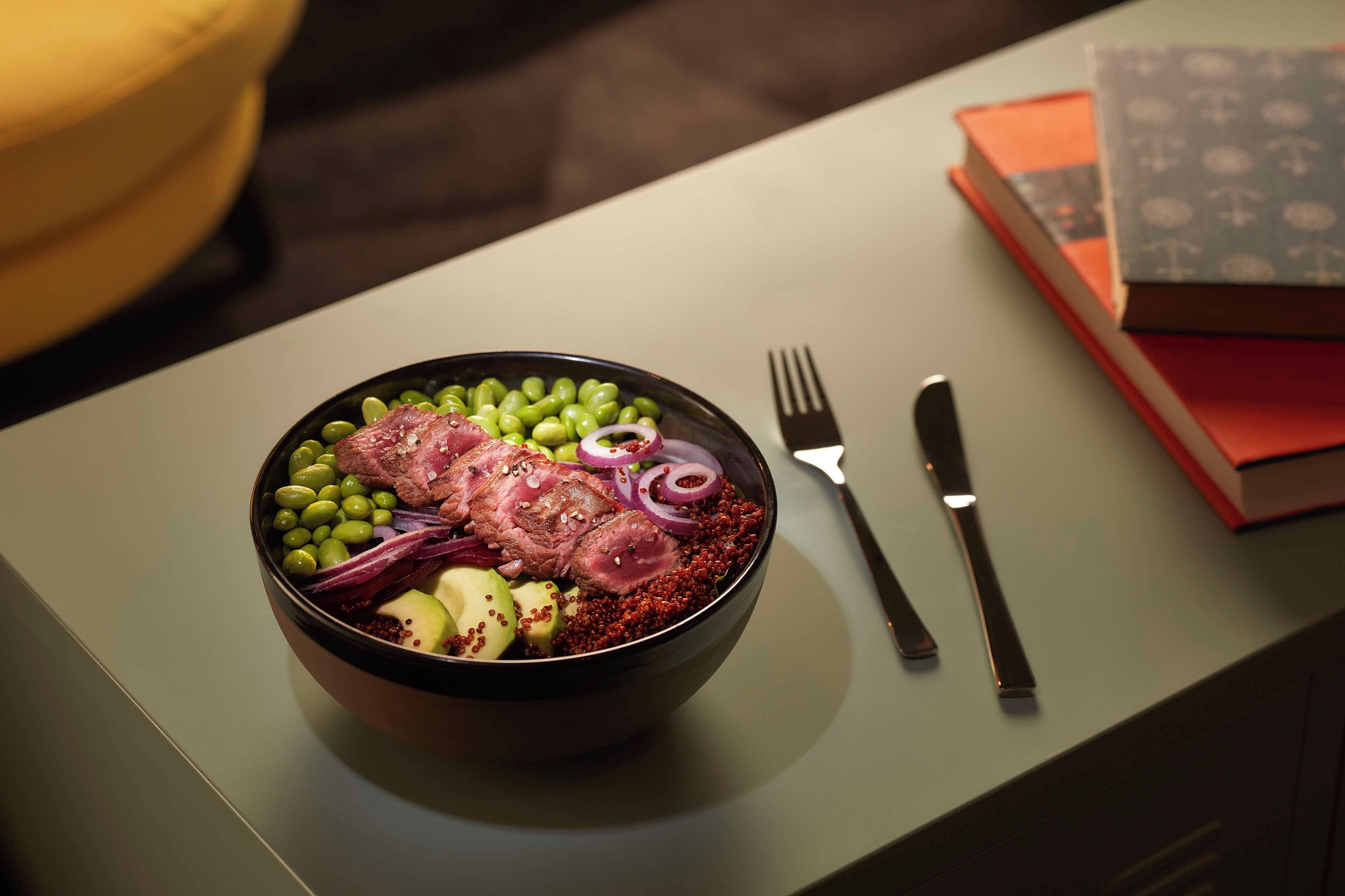 Eine Schüssel mit Essen, darunter Fleisch, grüne Bohnen und rote Zwiebeln, steht auf einem Tisch. Eine Gabel und ein Messer liegen daneben.