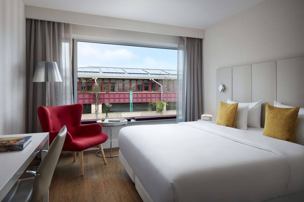 Ein Hotelzimmer mit einem großen Bett, einem roten Sessel, einem Schreibtisch und einem Fenster mit Blick auf ein Gebäude und einen Zug.
