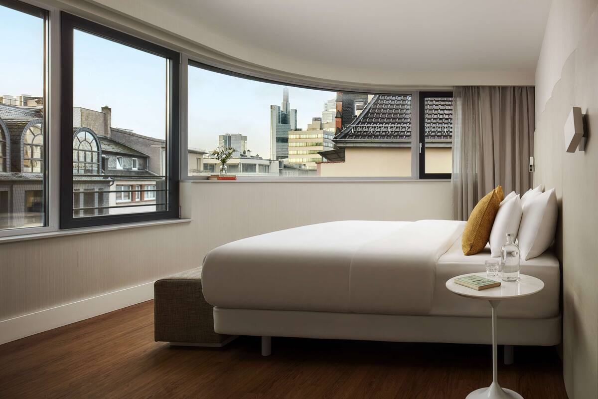 Ein modernes Hotelzimmer mit großem Fenster und Blick auf die Stadt, einem ordentlich bezogenen Bett, einem kleinen runden Tisch mit Buch und Glas sowie einer gemütlichen Sitzecke.