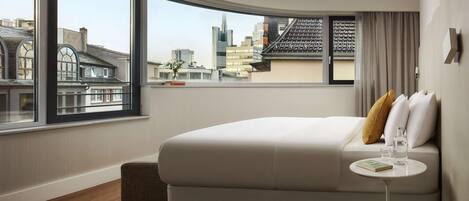 A modern hotel room with a large window offering a city view, a neatly made bed, a small round table with a book and glass, and a cozy seating area.
