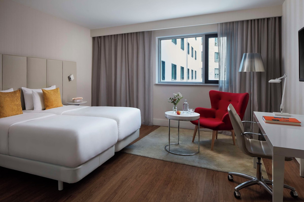Habitación de hotel con dos camas, un escritorio, una silla, una lámpara y vistas a edificios a través de la ventana.