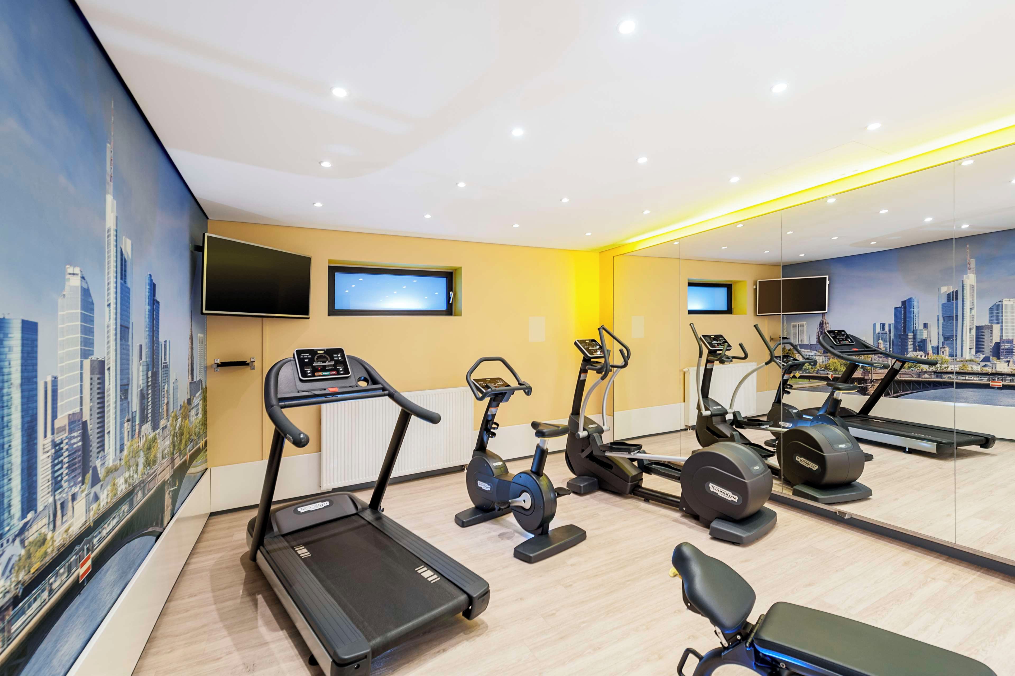 Un gimnasio con cintas de correr, bicicletas estáticas y elípticas, que presenta murales con vistas a la ciudad y múltiples pantallas de televisión.