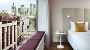 Une chambre d’hôtel dotée d’une grande fenêtre donnant sur la ville, un lit blanc agrémenté d’un coussin décoratif doré, une petite table ronde avec un livre et un fruit, et un banc placé le long de la fenêtre.