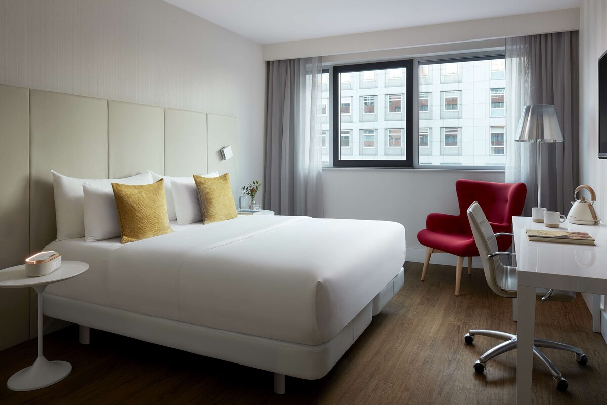 Een moderne hotelkamer met een groot bed, een bureau, een rode stoel en een raam met uitzicht op de stad.