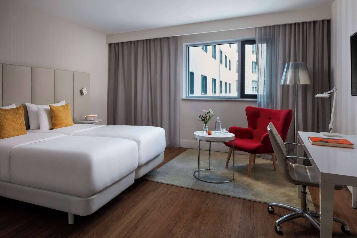 Ein Hotelzimmer mit zwei Betten, einem Schreibtisch, einem Stuhl, einer Lampe und Blick auf Gebäude durch das Fenster.