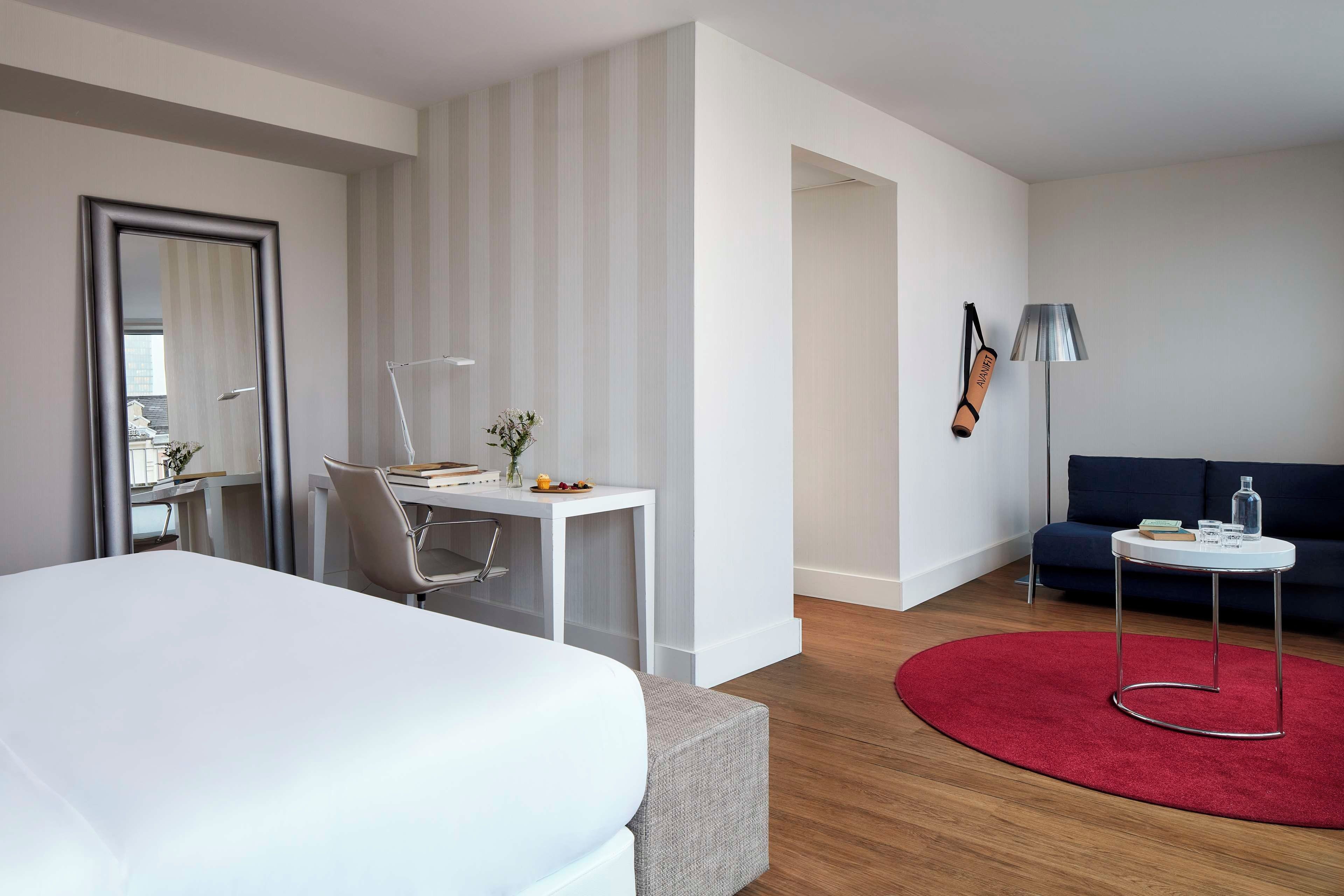 A hotel room with a bed, a desk, a chair, a small table, a sofa, a red rug, and a lamp.