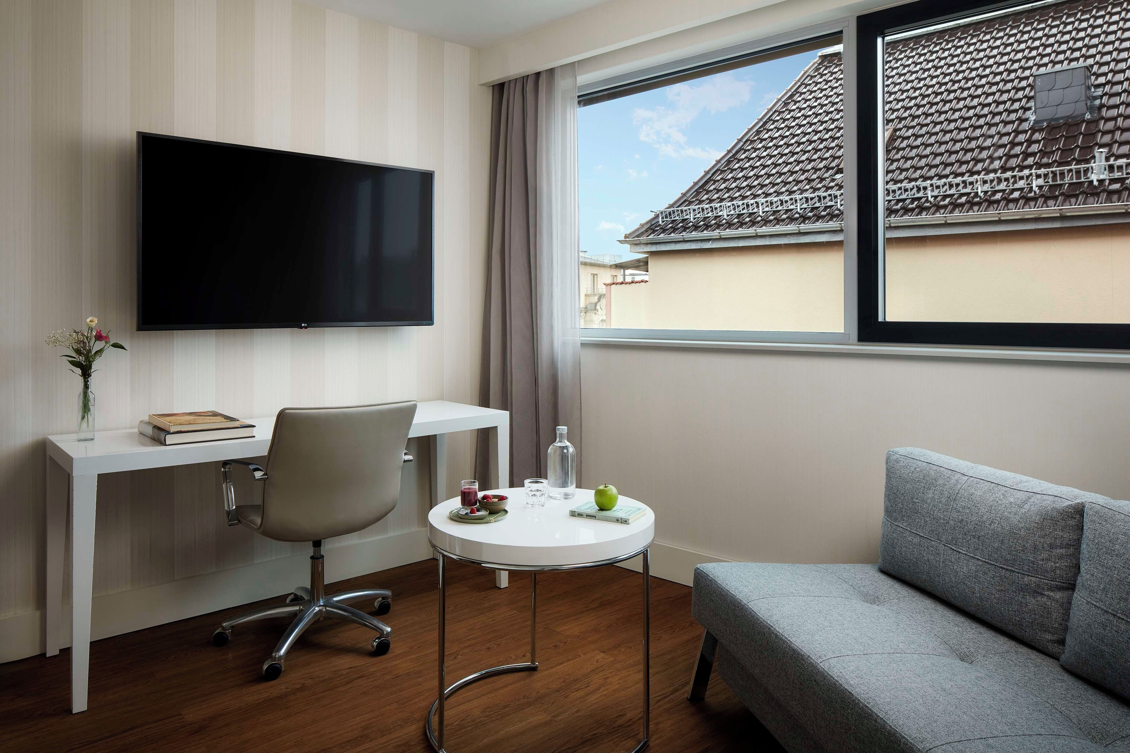 A modern hotel room with a flat-screen TV, a desk with a chair, a small round table with a glass of water, a bowl of fruit, and a grey sofa.