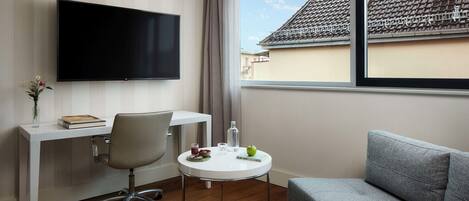 Una habitación de hotel moderna con un televisor de pantalla plana, un escritorio con una silla, una pequeña mesa redonda con un vaso de agua, un cuenco de fruta y un sofá gris.