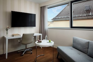 Une chambre d’hôtel moderne équipée d’un téléviseur à écran plat, d’un bureau avec une chaise, d’une petite table ronde avec un verre d’eau, d’un bol de fruits et d’un canapé gris.
