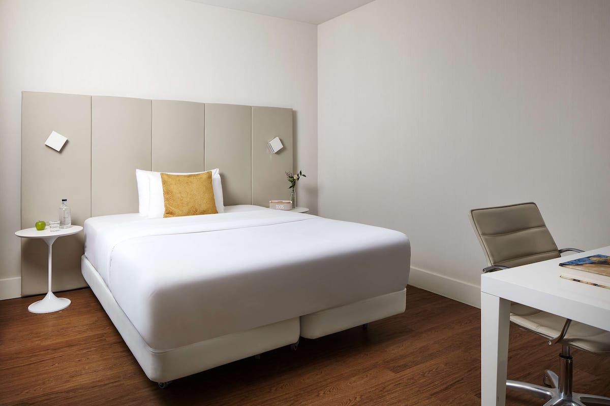 Ein Hotelzimmer mit einem großen Bett, einem kleinen Nachttisch mit einem Glas, einem grünen Apfel und einer Vase mit Blumen. Es gibt einen weißen Schreibtisch mit einem Stuhl und einem Notizbuch.