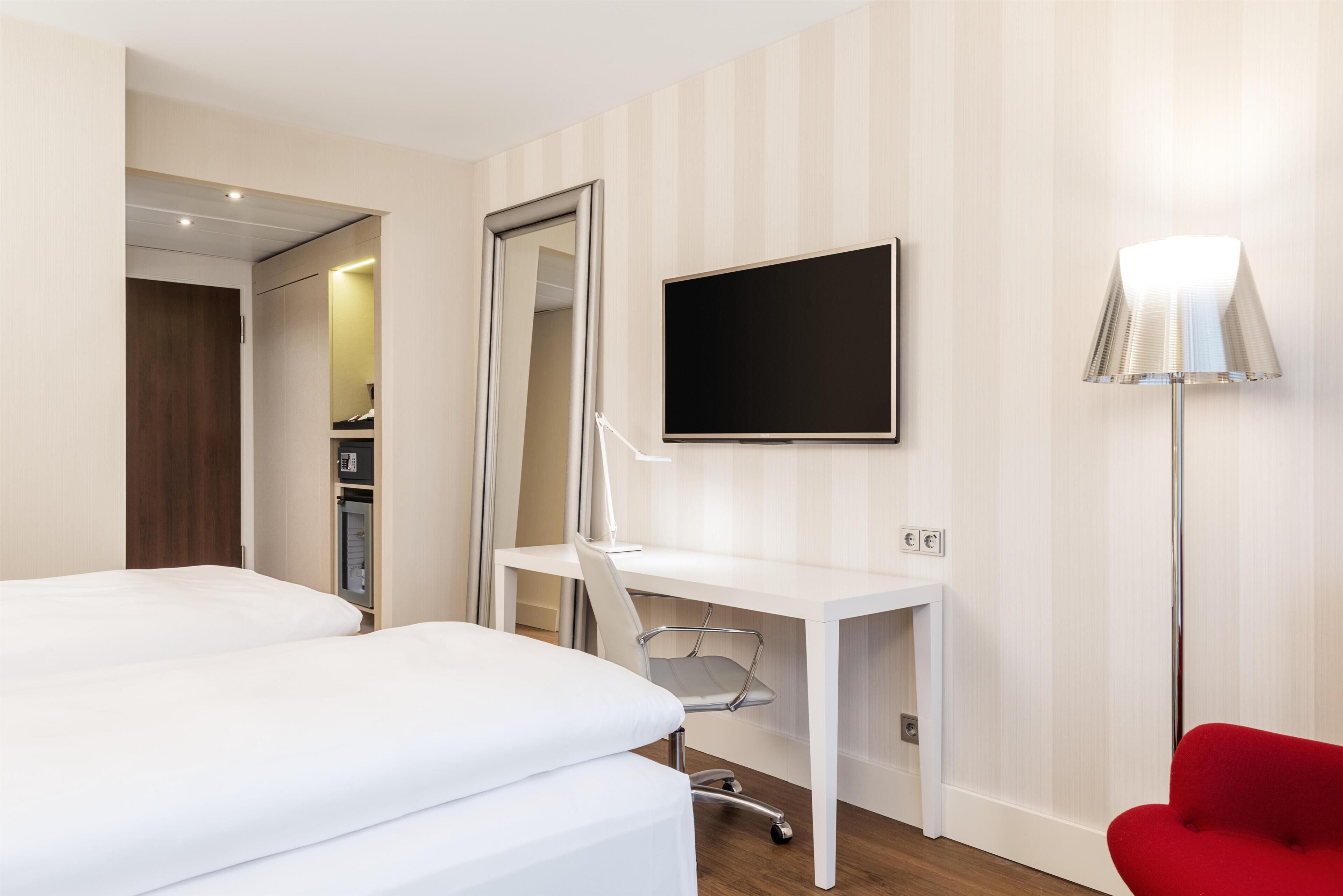 A hotel room with a bed, a desk with a chair, a television, and a red chair.