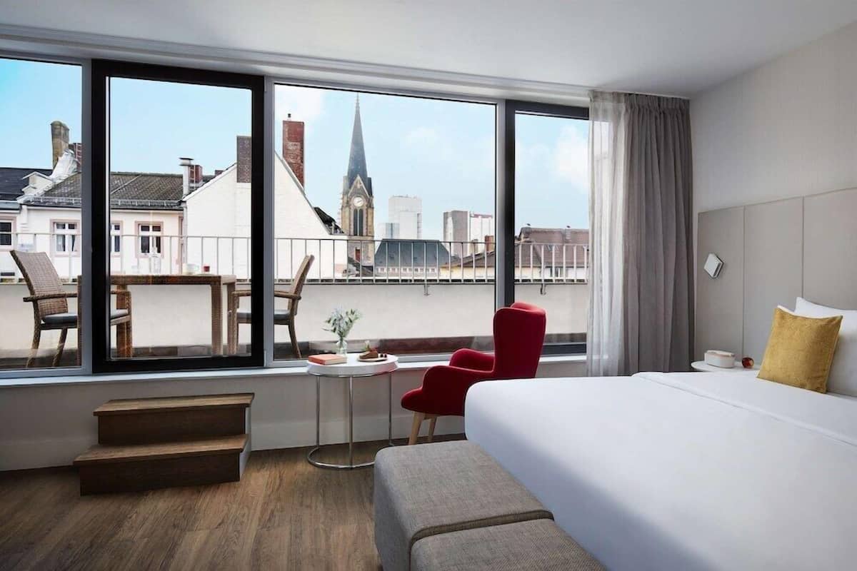 Ein Hotelzimmer mit großem Fenster, einem Bett, einem roten Sessel, einem kleinen Tisch und einer Bank.