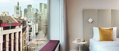 A hotel room with a city view, a white sofa, a small round table, and a window with a view of buildings and a street.