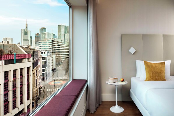 A hotel room with a city view, a white sofa, a small round table, and a window with a view of buildings and a street.