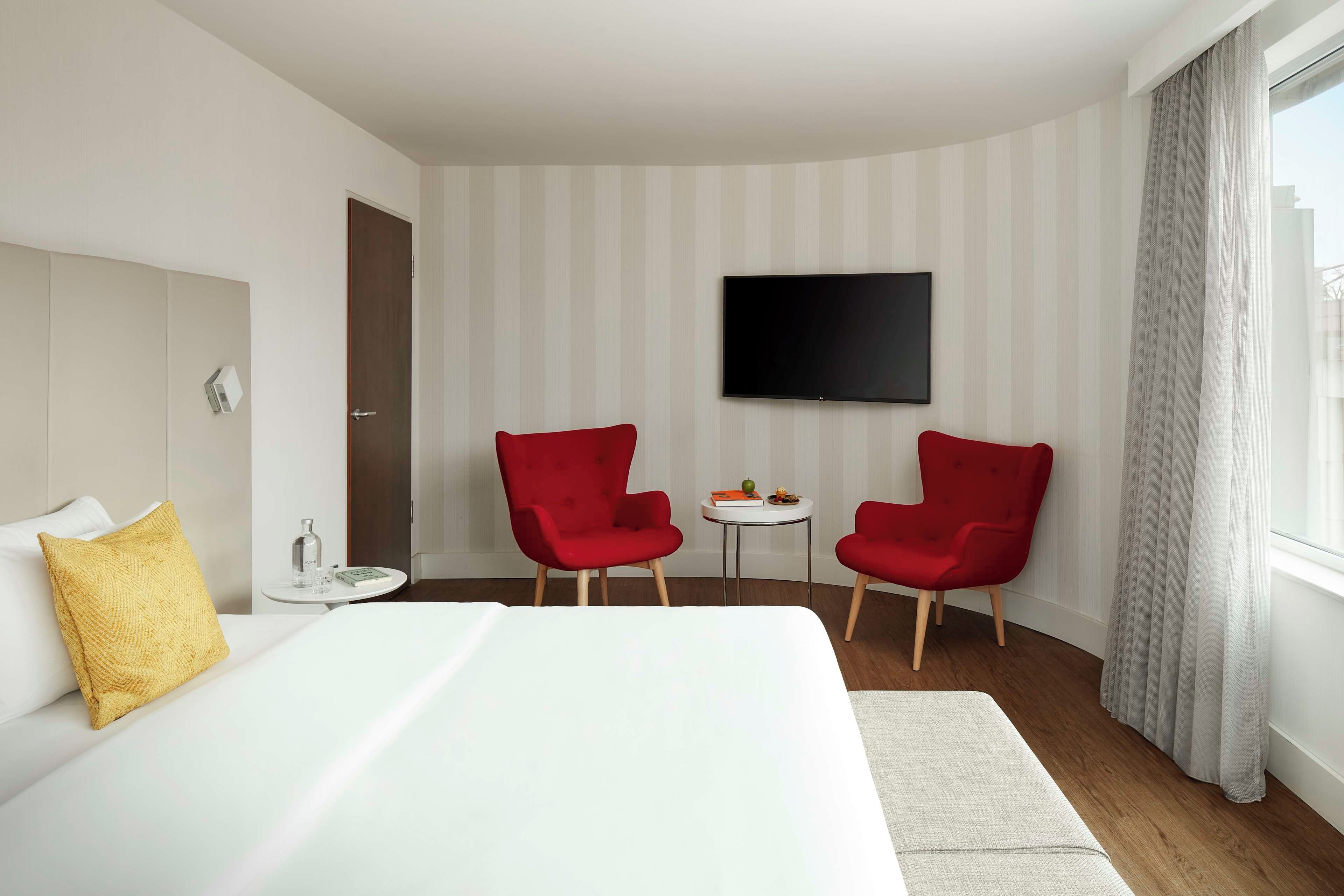 A hotel room with a bed, two red chairs, a small table with a book and a bottle, a TV on the wall, and a window with curtains.