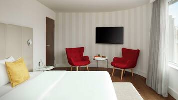 A hotel room with a bed, two red chairs, a small table with a book and a bottle, a TV on the wall, and a window with curtains.