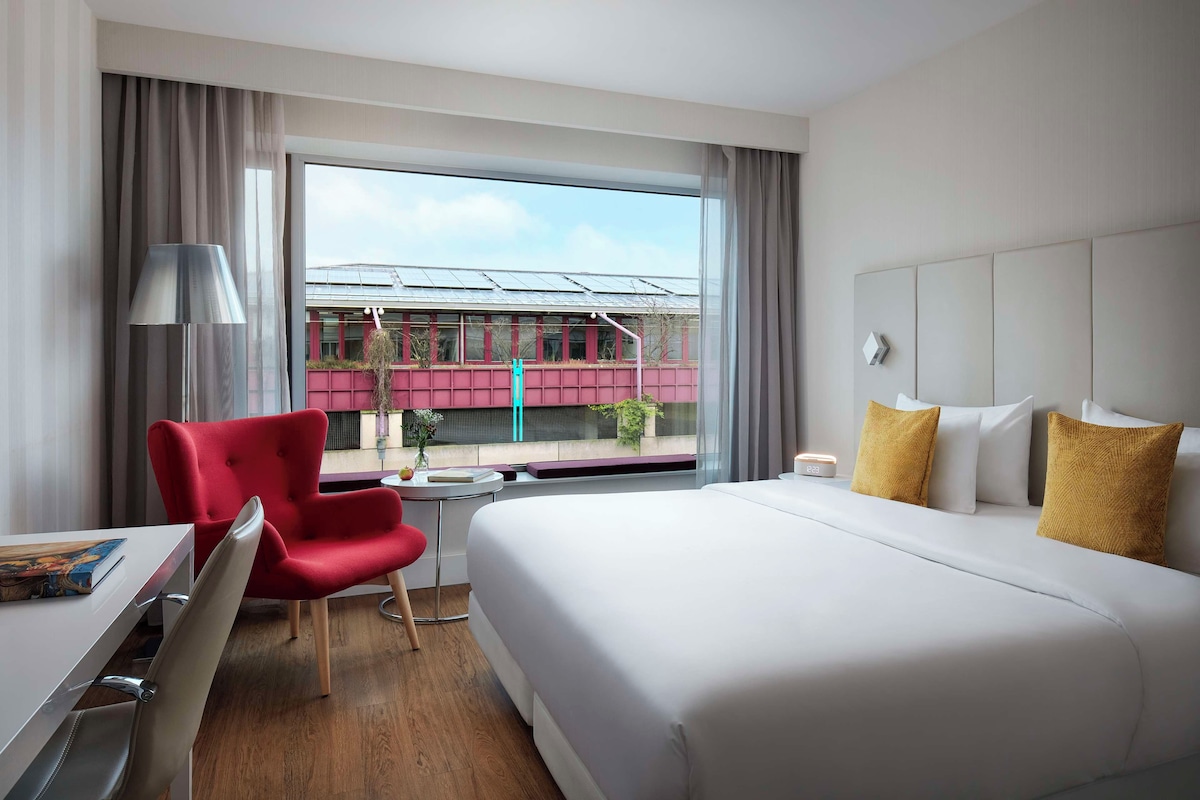 Habitación de hotel con una cama grande, una silla roja, un escritorio y una ventana que ofrece vistas a un edificio y un tren.