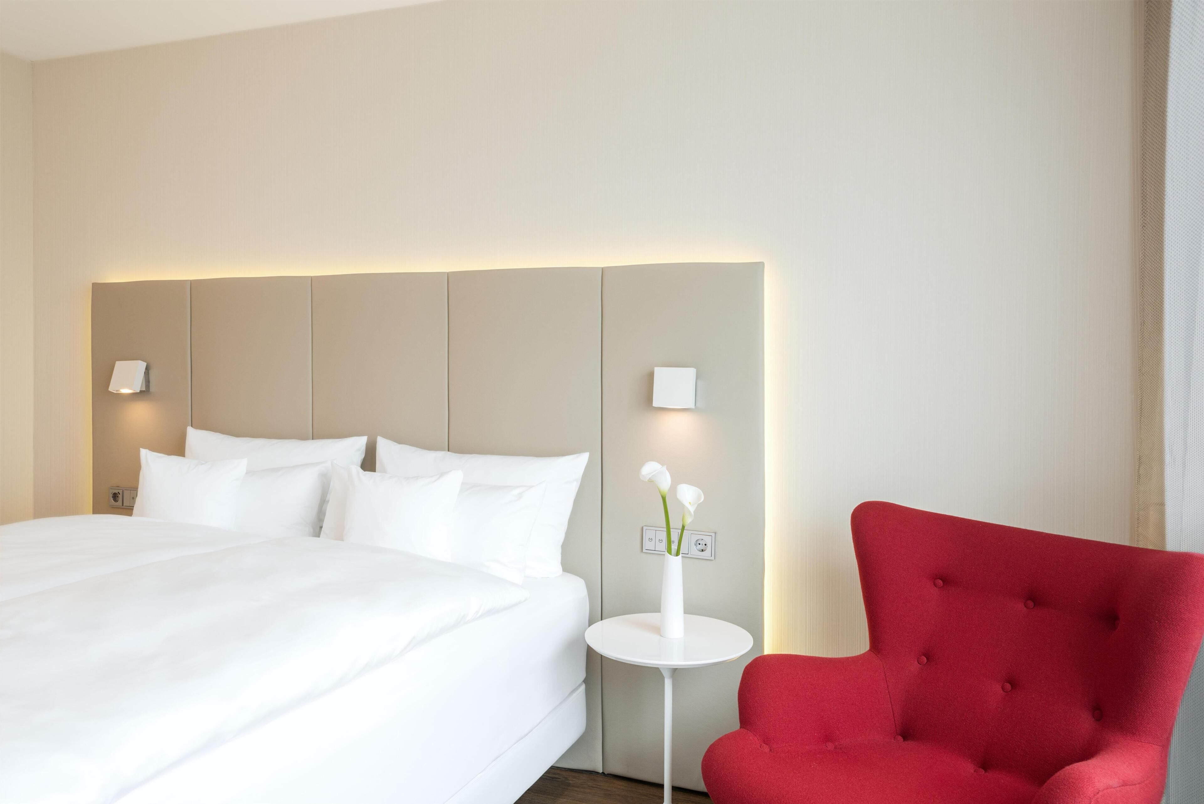 Habitación de hotel con una cama grande, un sillón rojo y una mesita con un jarrón de flores.