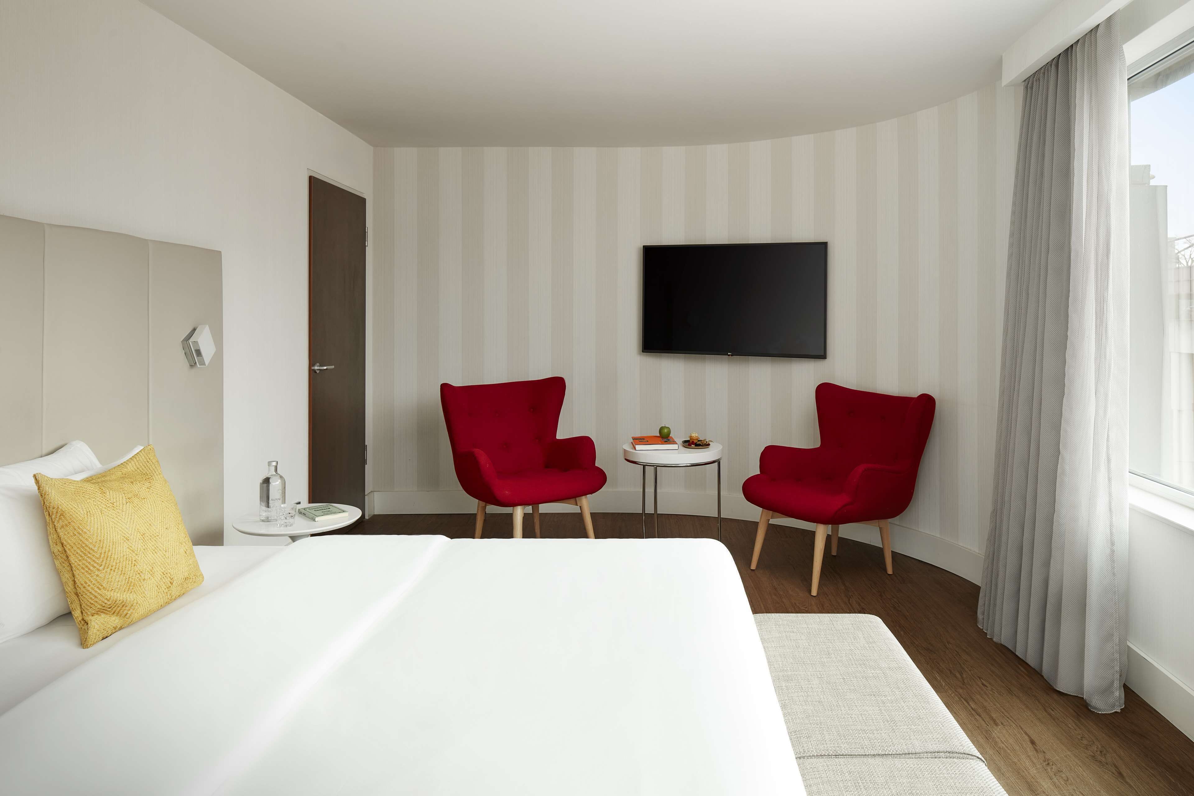 Una habitación de hotel con una cama grande, dos sillas rojas, una mesita con una botella y un libro, un televisor de pantalla plana en la pared y una ventana con cortinas.