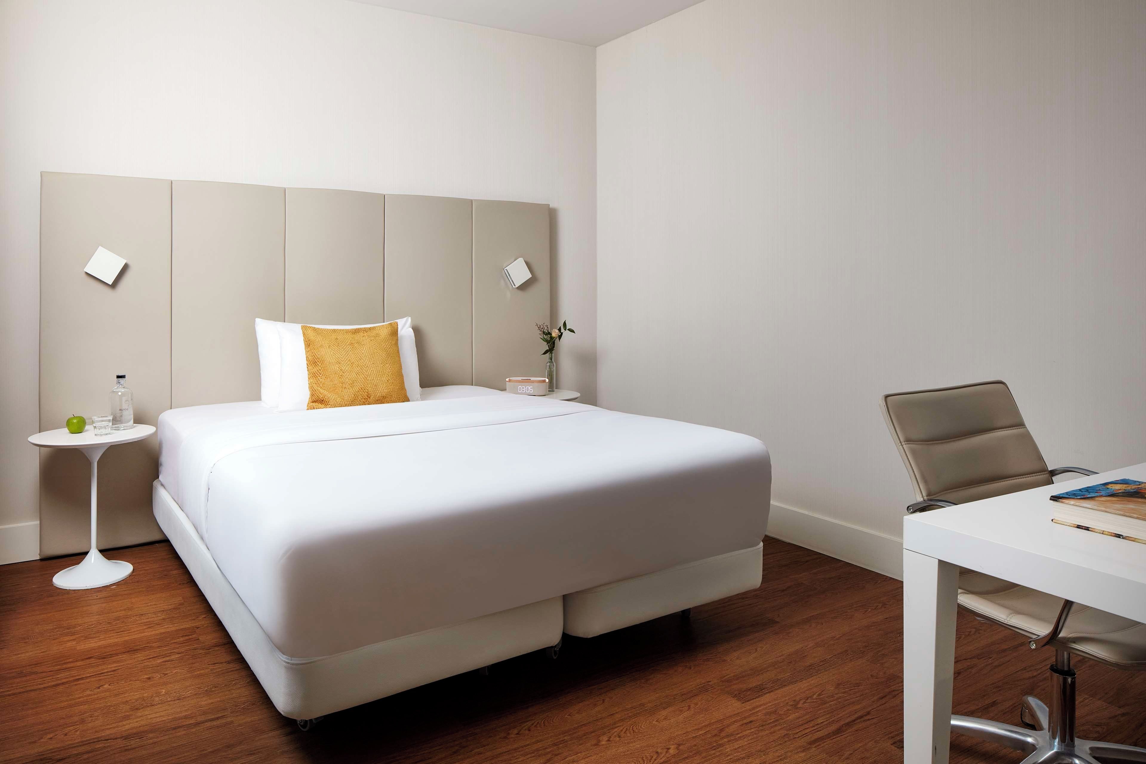 Habitación de hotel con una cama grande, una mesita de noche con un vaso, una manzana verde y un jarrón con flores. Hay un escritorio blanco con una silla y un cuaderno.