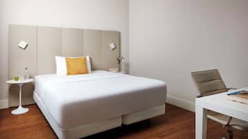 Ein Hotelzimmer mit einem großen Bett, einem kleinen Nachttisch mit einem Glas, einem grünen Apfel und einer Vase mit Blumen. Es gibt einen weißen Schreibtisch mit einem Stuhl und einem Notizbuch.