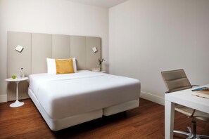 A hotel room with a bed, a small bedside table, a chair, a desk, and a clock.