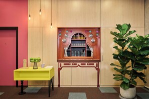 Una habitación con un armario de madera con puertas de cristal, una mesa amarilla con un jarrón de manzanas verdes y una planta grande en maceta.