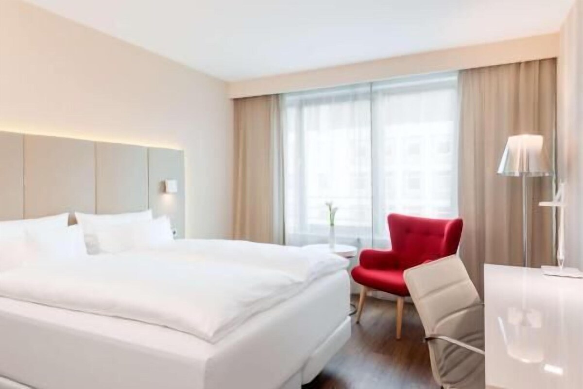 Une chambre d’hôtel avec un grand lit, un fauteuil rouge, un bureau et une fenêtre avec des rideaux.