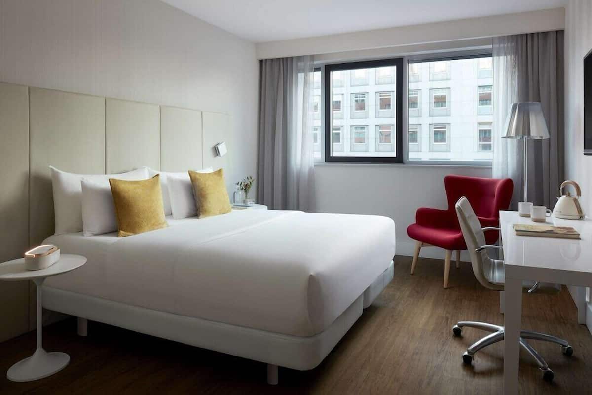 Ein Hotelzimmer mit Bett, Schreibtisch, Stuhl, Lampe und Blick auf Gebäude.