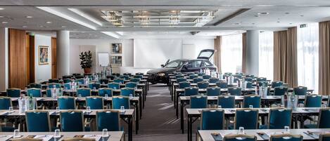 A conference room with rows of tables and chairs, a large black car parked in the center, and a potted plant in the corner.