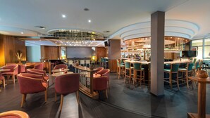 A modern bar area with round tables and bar stools, a television, and a variety of bottles and glasses on the shelves. - Congress Hotel Weimar by Mercure (Weimar)