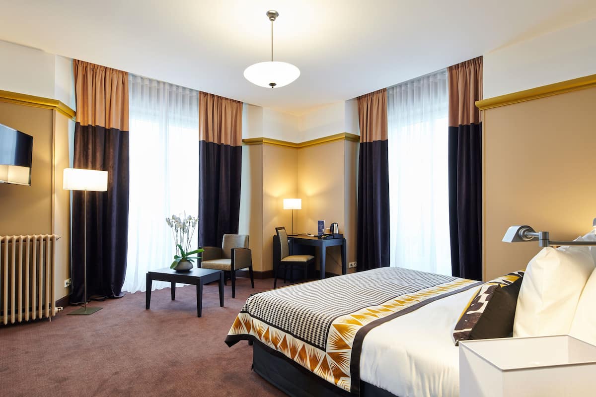 A hotel room with a large bed, a desk, a chair, a lamp, and a radiator.