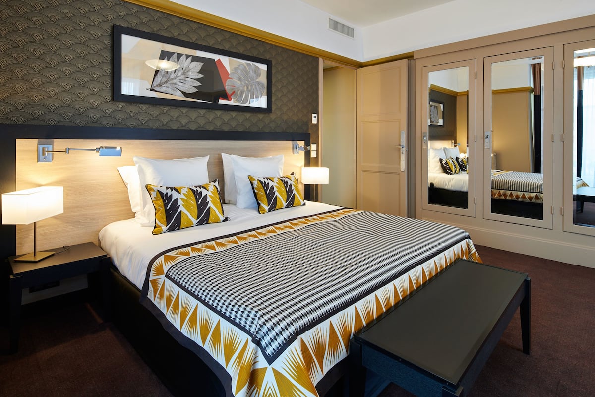 A hotel room with a large bed, bedside lamps, a bench, a wardrobe, and a framed artwork on the wall.