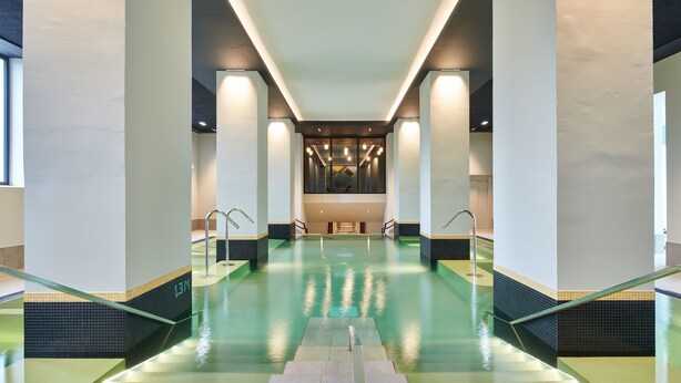 Une piscine intérieure moderne, avec une eau cristalline, entourée de murs et de colonnes blancs, et un escalier central menant à un espace détente.