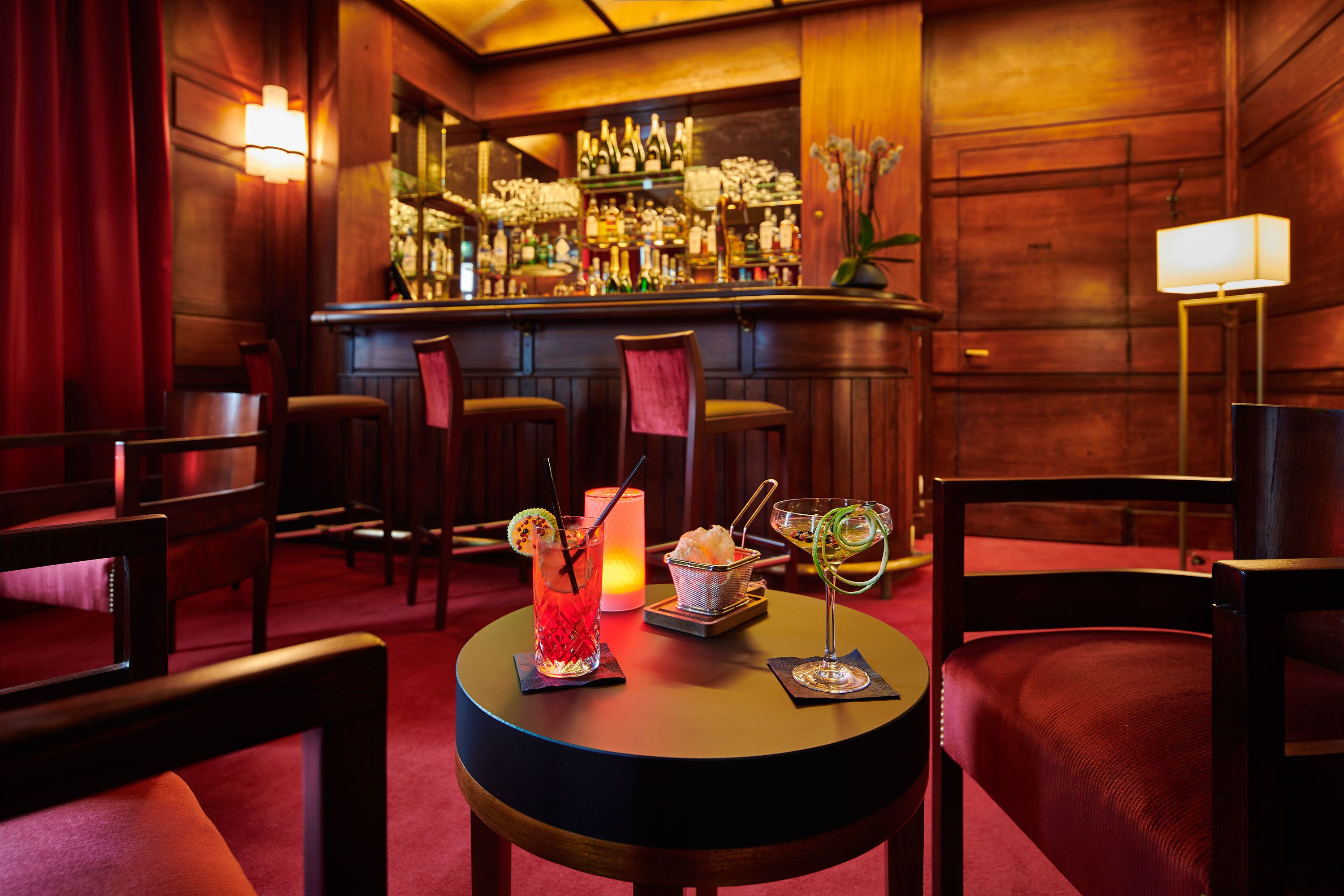 A bar area with wooden paneling, a well-stocked bar, and a table with two drinks and a candle.