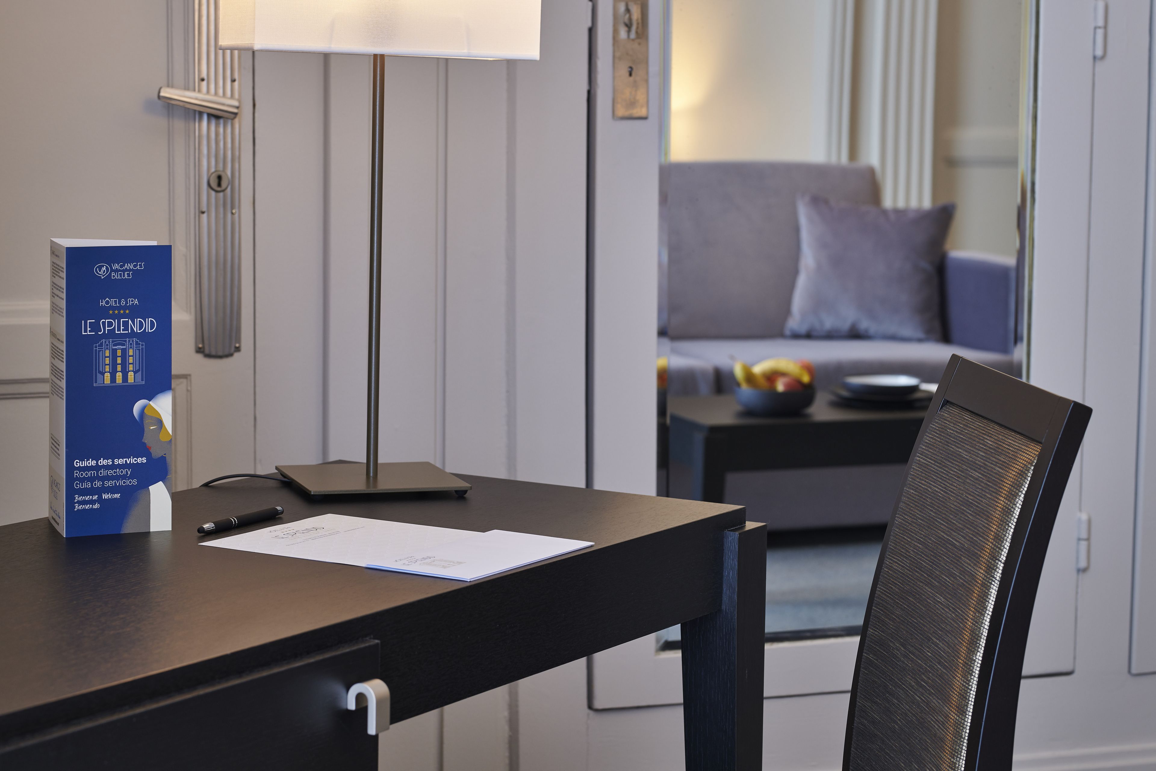 A hotel room with a desk, a lamp, a chair, a sofa, and a small table with a bowl of fruit.