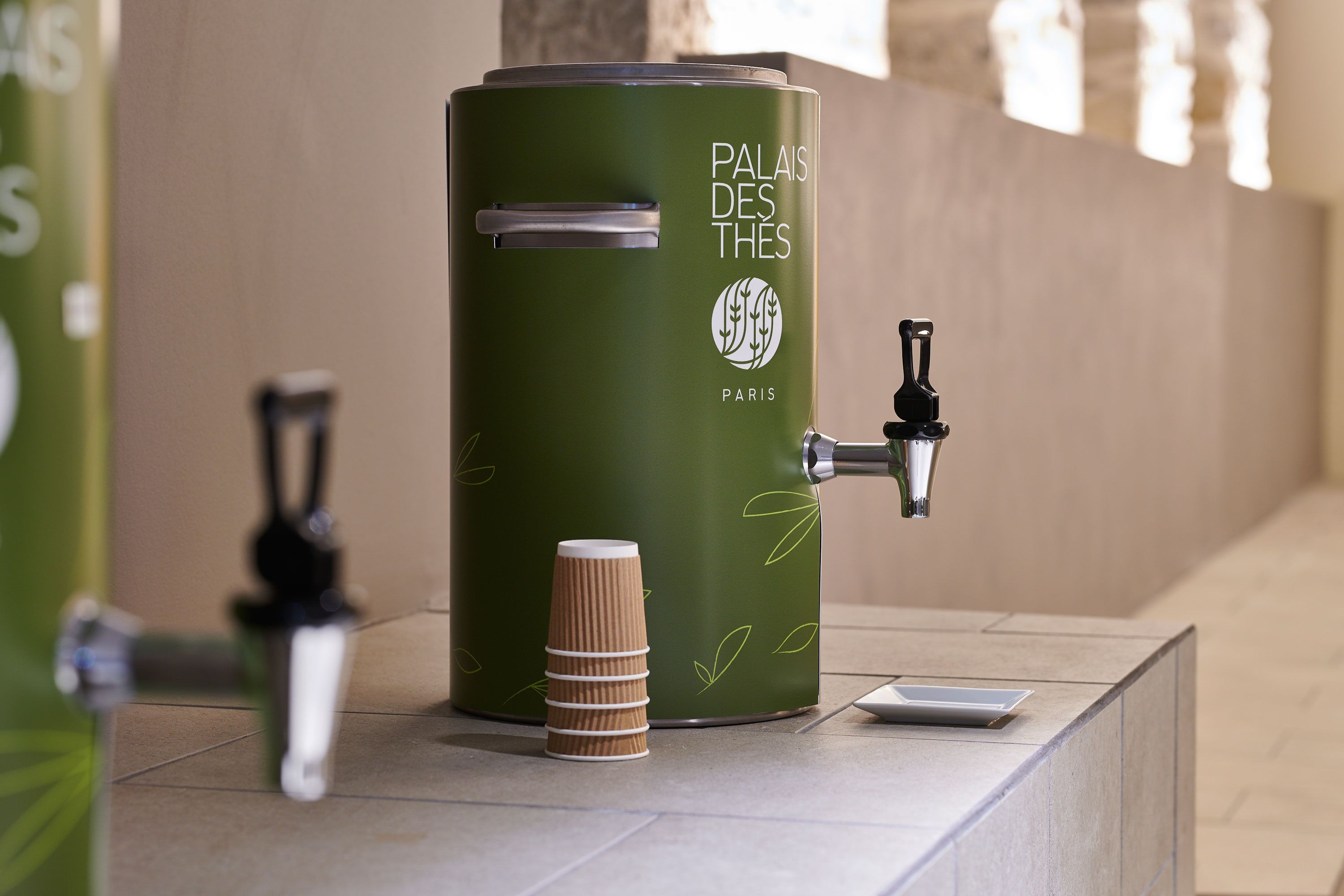 A green trash can with a tap and a stack of paper cups, labeled "Palais des Thés Paris".