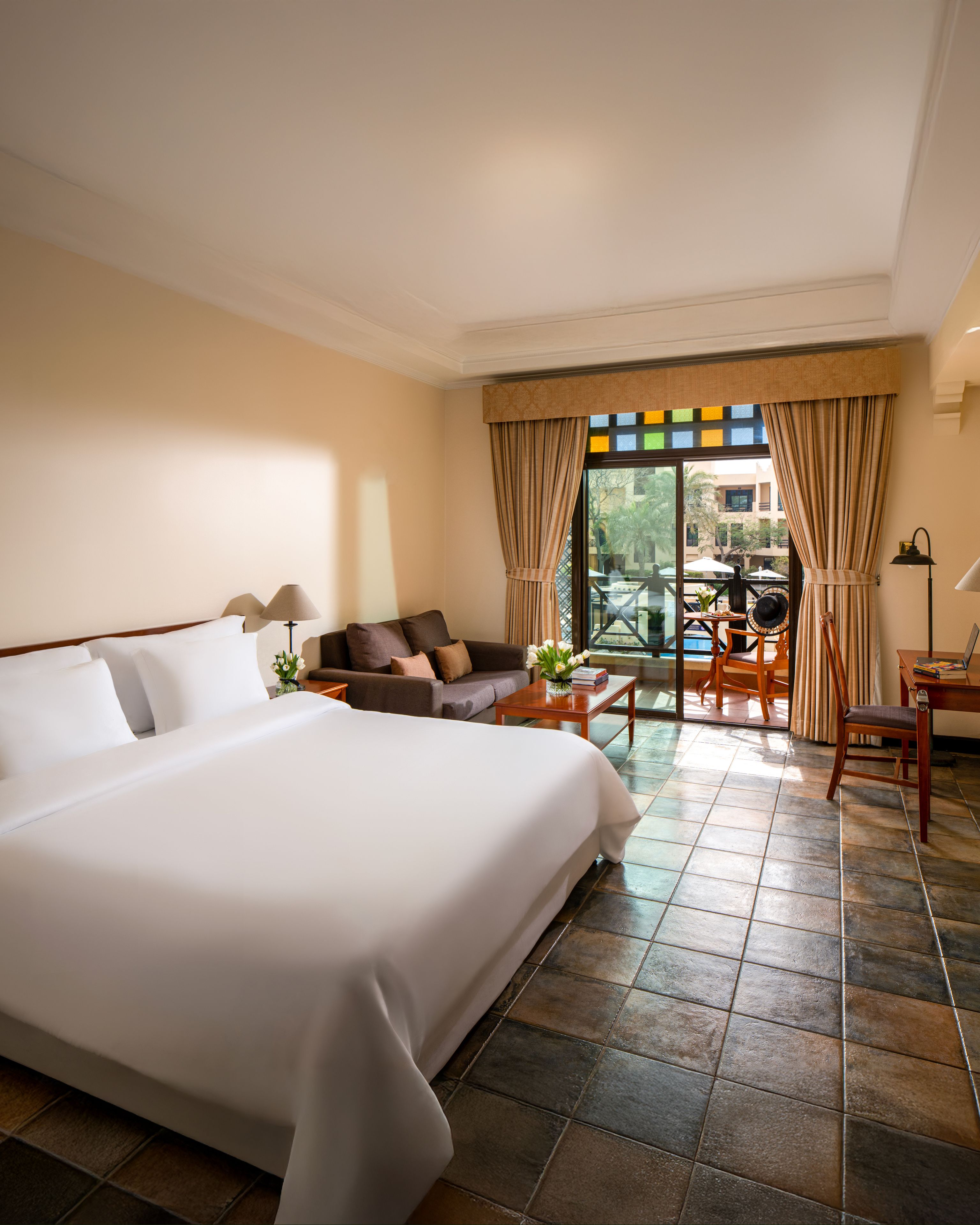 A hotel room with a large bed, a sofa, a small table, and a balcony area with a chair and umbrella.