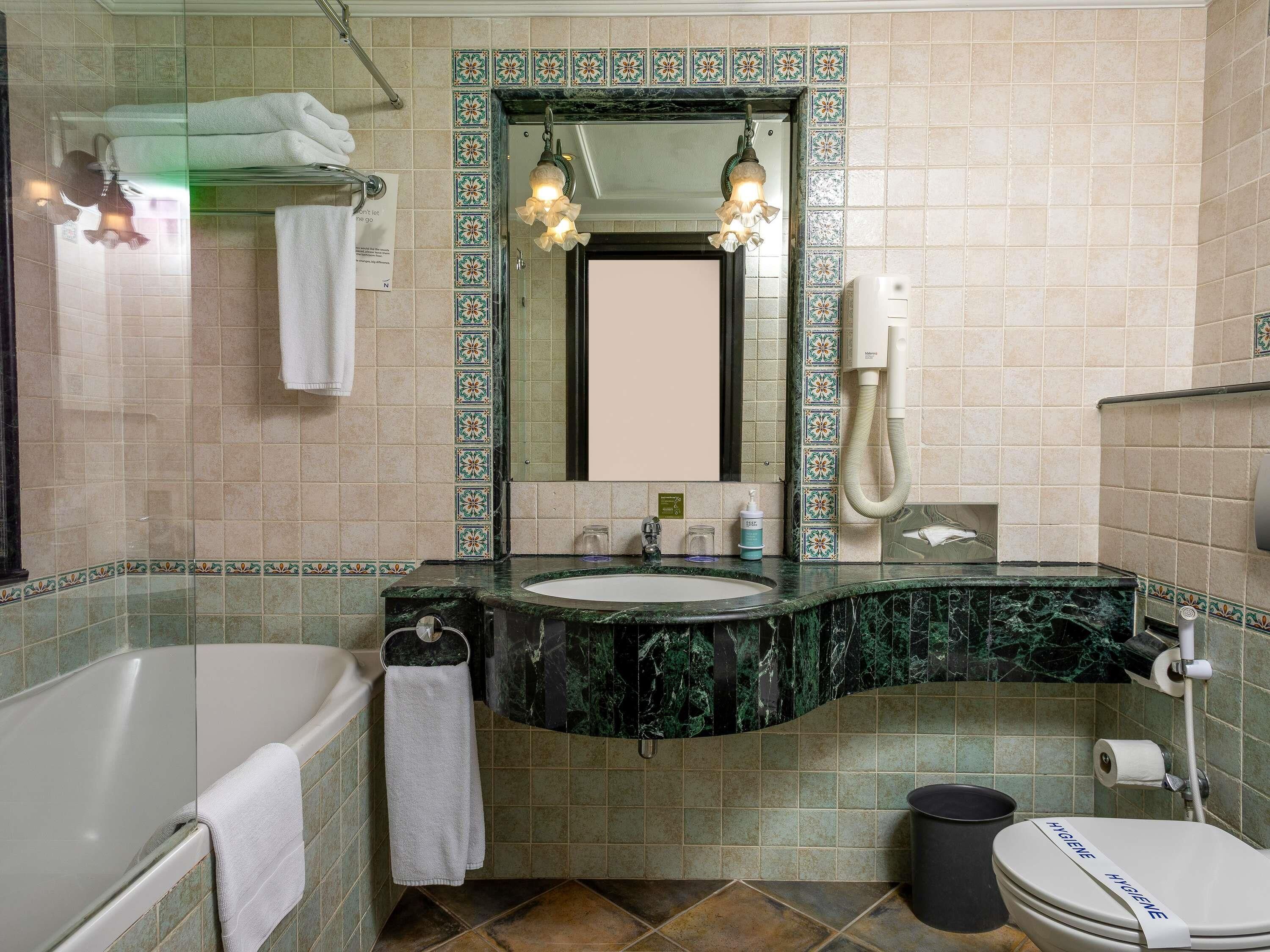 A bathroom with a bathtub, a sink with a marble countertop, a mirror with a decorative border, a towel rack, and a toilet with a hygiene sign.