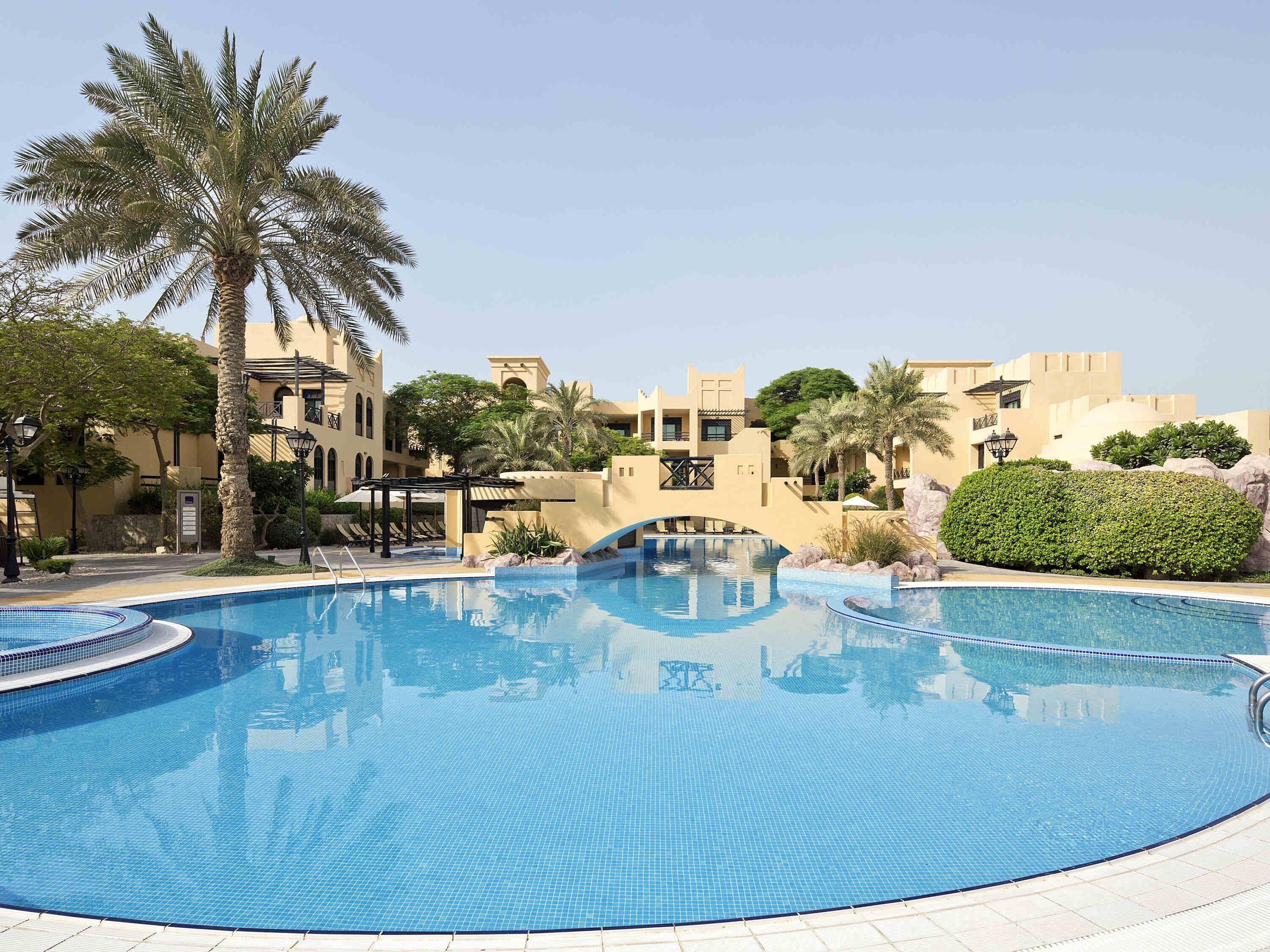 A swimming pool with a bridge, palm trees, and buildings in the background.