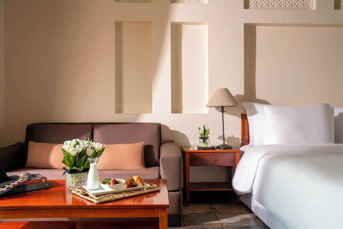 A hotel room with a sofa, a bedside table with a lamp, a bed, and a tray with food and flowers.