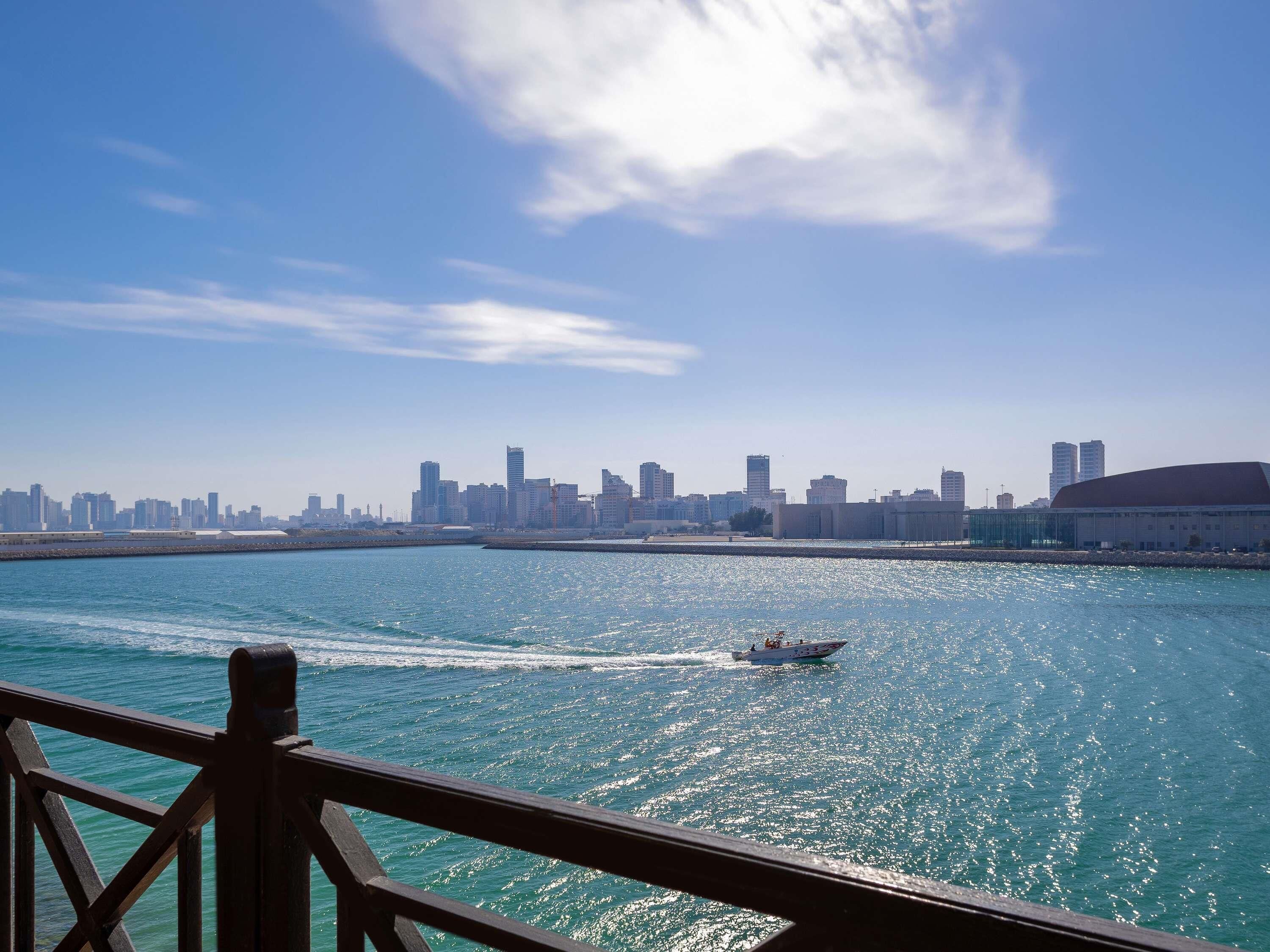 A waterfront cityscape with a boat on the water, a clear sky, and a distant view of a city skyline.