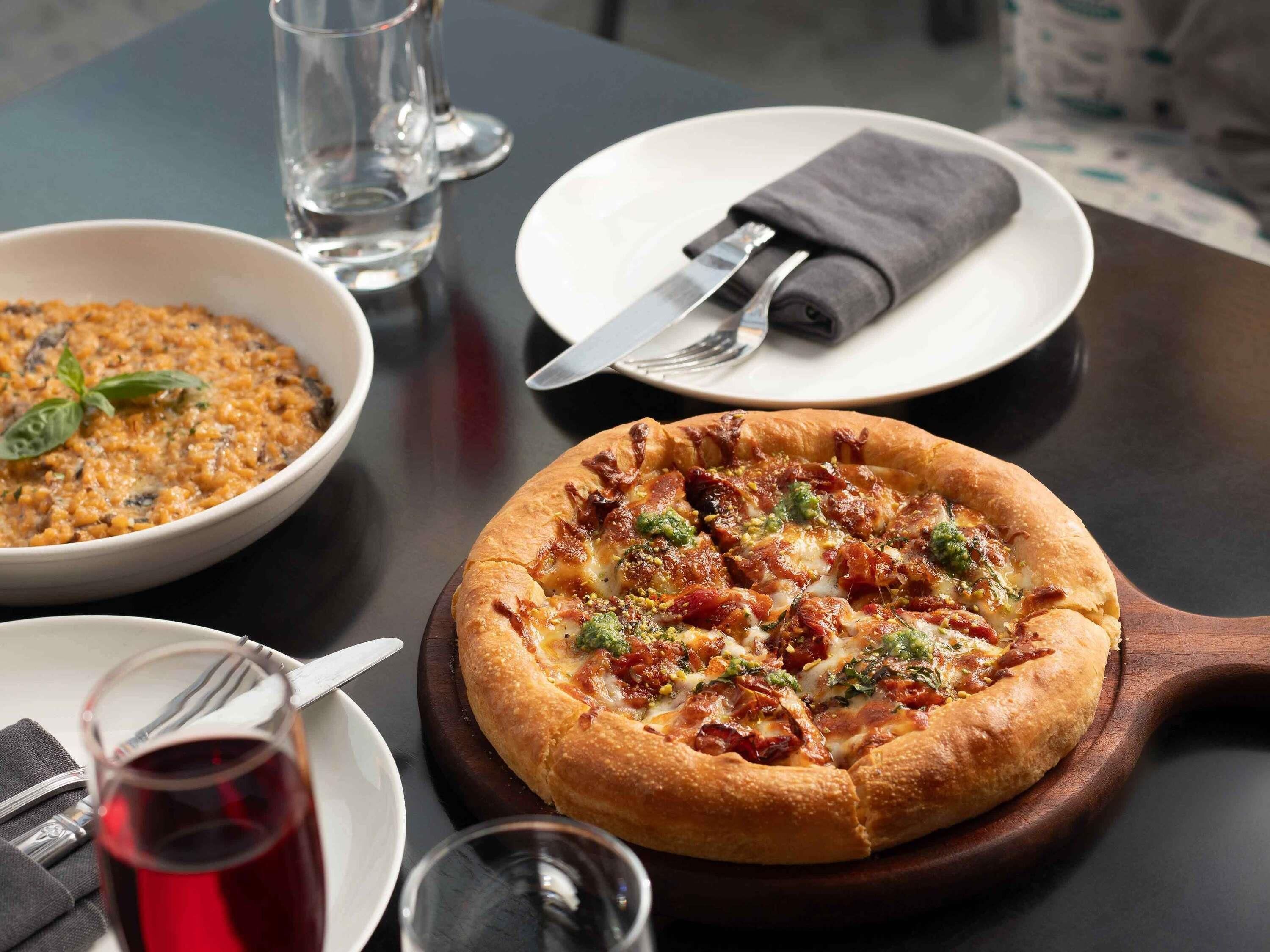 Une pizza sur une planche en bois, un bol de nourriture, un verre de boisson rouge et une assiette avec un couteau et une serviette sur une table.