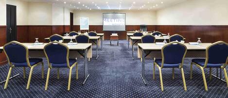 Un salón de conferencias con filas de mesas y sillas, una pantalla de proyector y suelo alfombrado.