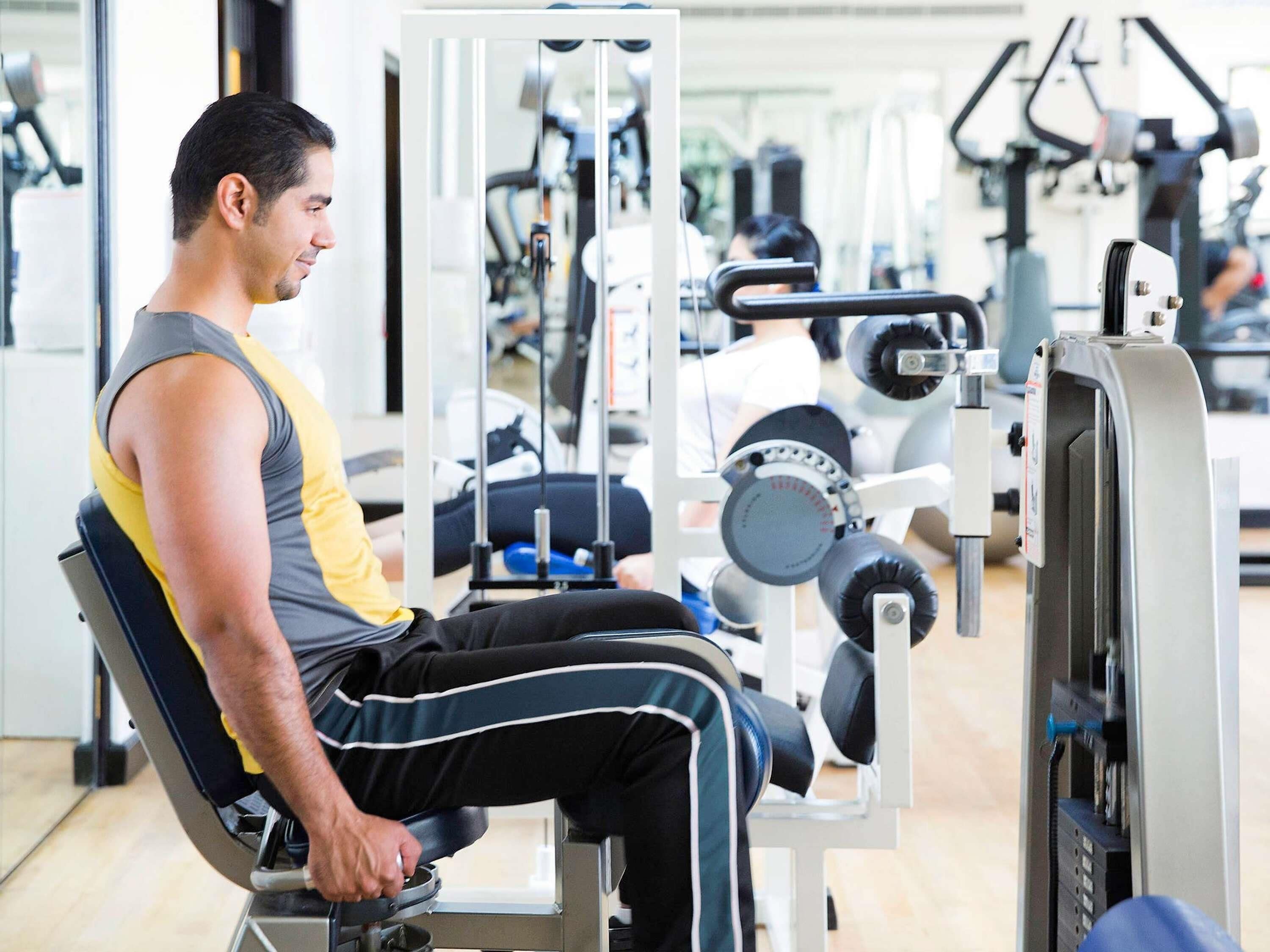 A gym with exercise equipment, a seated person using a machine, and a window in the background.