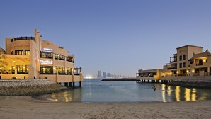 A waterfront hotel with a sandy beach, a city skyline in the background, and a calm water body. - Novotel Bahrain Al Dana Resort (Manama)